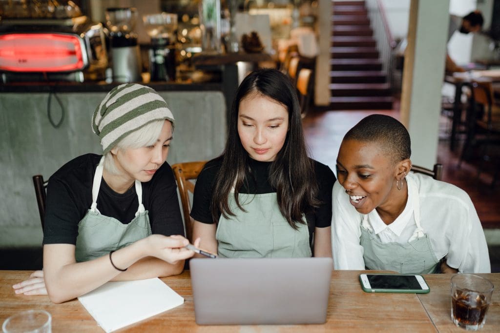 Three people sit in a coffee shop with green aprons on looking at a laptop